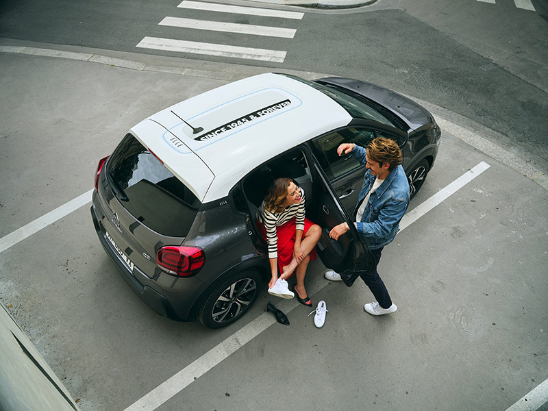 Un homme plaisante en s'appuyant sur la portière de la Citroën C3, tandis qu'une femme, assise à l'arrière de la voiture, échange ses talons hauts pour des baskets confortables.